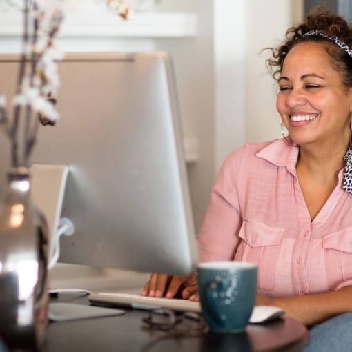 Person sits at desktop computer with coffee mug and decorative flowers in a vase.