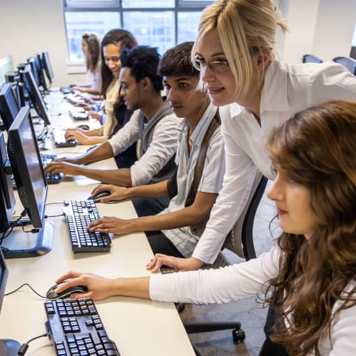 Students sit in front of computers in a classroom while being instructed by a teacher. 