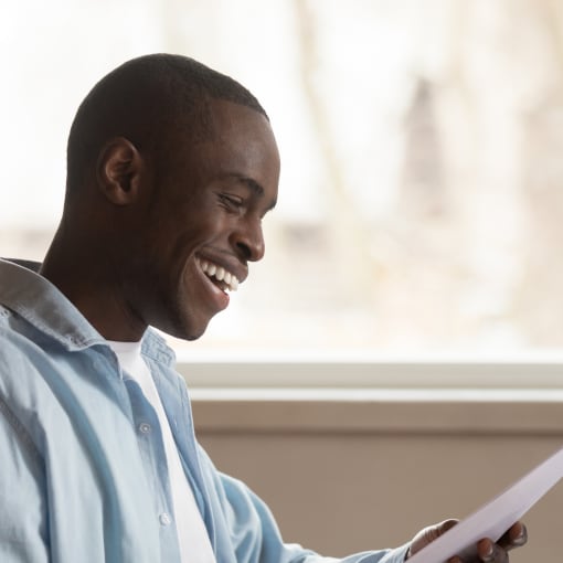 Prospective student smiling, sitting at desk and looking at a piece of paper as they prepare to enroll for school on a laptop.