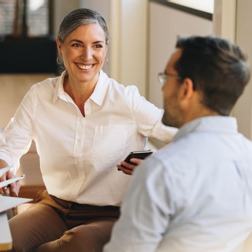 Two people smile and talk as they sit at a table. There is a laptop and a book on the table. 