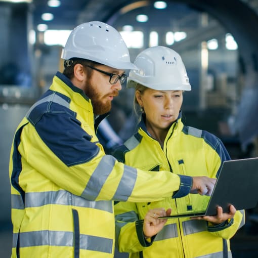 Two employees of a manufacturing company discussing information. One is holding a laptop, the other one is pointing at some data on the screen.