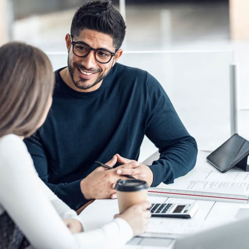 Two people converse while sitting at a small desk with a coffee cup, papers and a calculator on it.