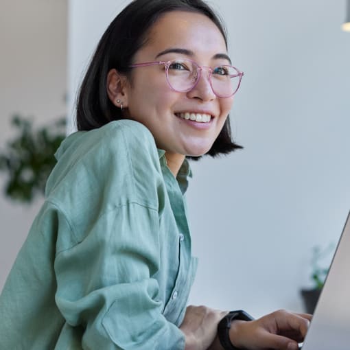 Person in glasses and a teal blouse smiles while working on a laptop.