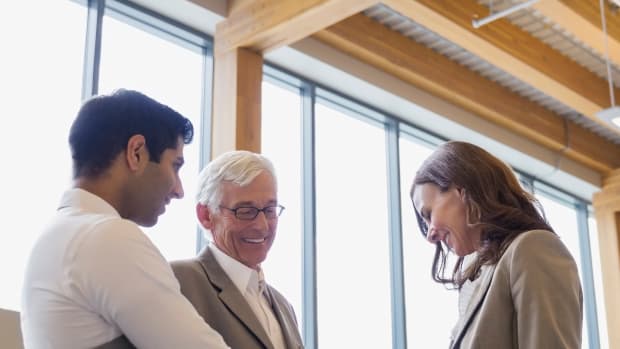 Three people smile and talk in an office. Two are holding clipboards. 