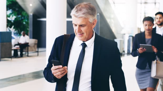 Person in suit walks down hallway while looking at cell phone.