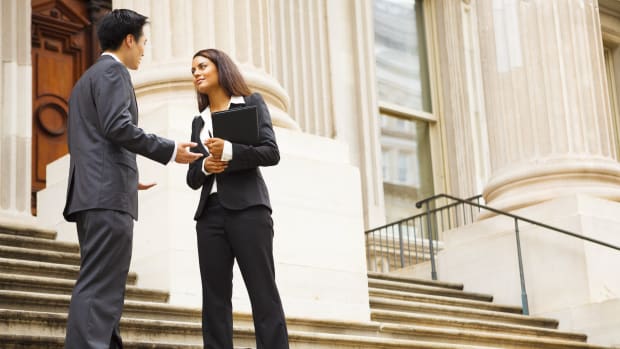 Two people standing on the stairs in front of a governmental building discussing a case. One is holding  files and a pen.