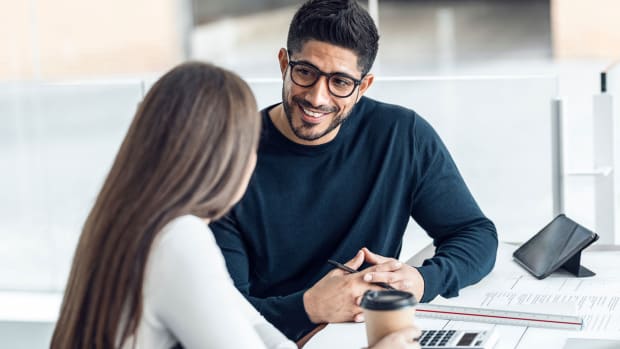 Two people converse while sitting at a small desk with a coffee cup, papers and a calculator on it.