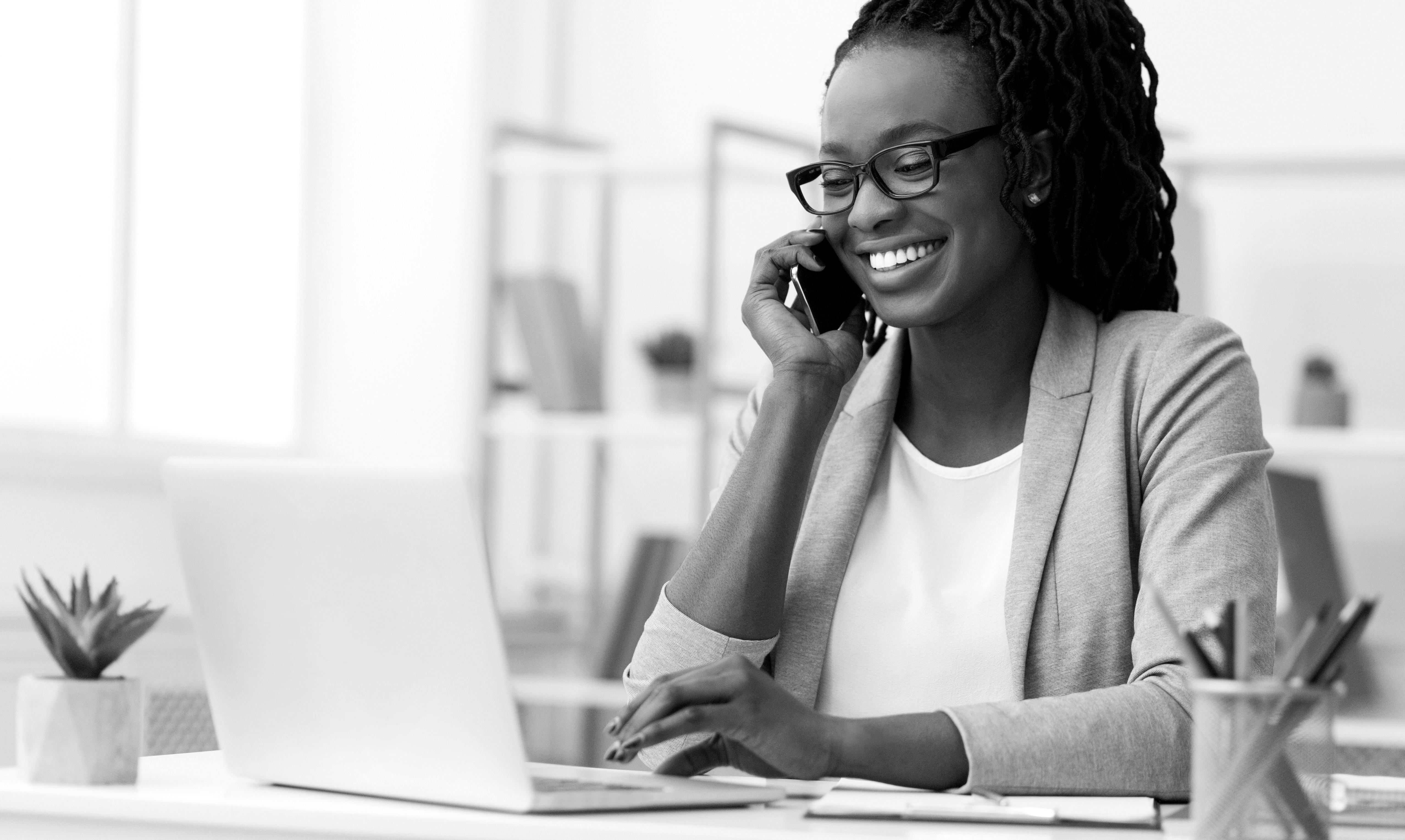 Black-and-white photo of a business professional sitting at a desk with a laptop, pens and small plant.