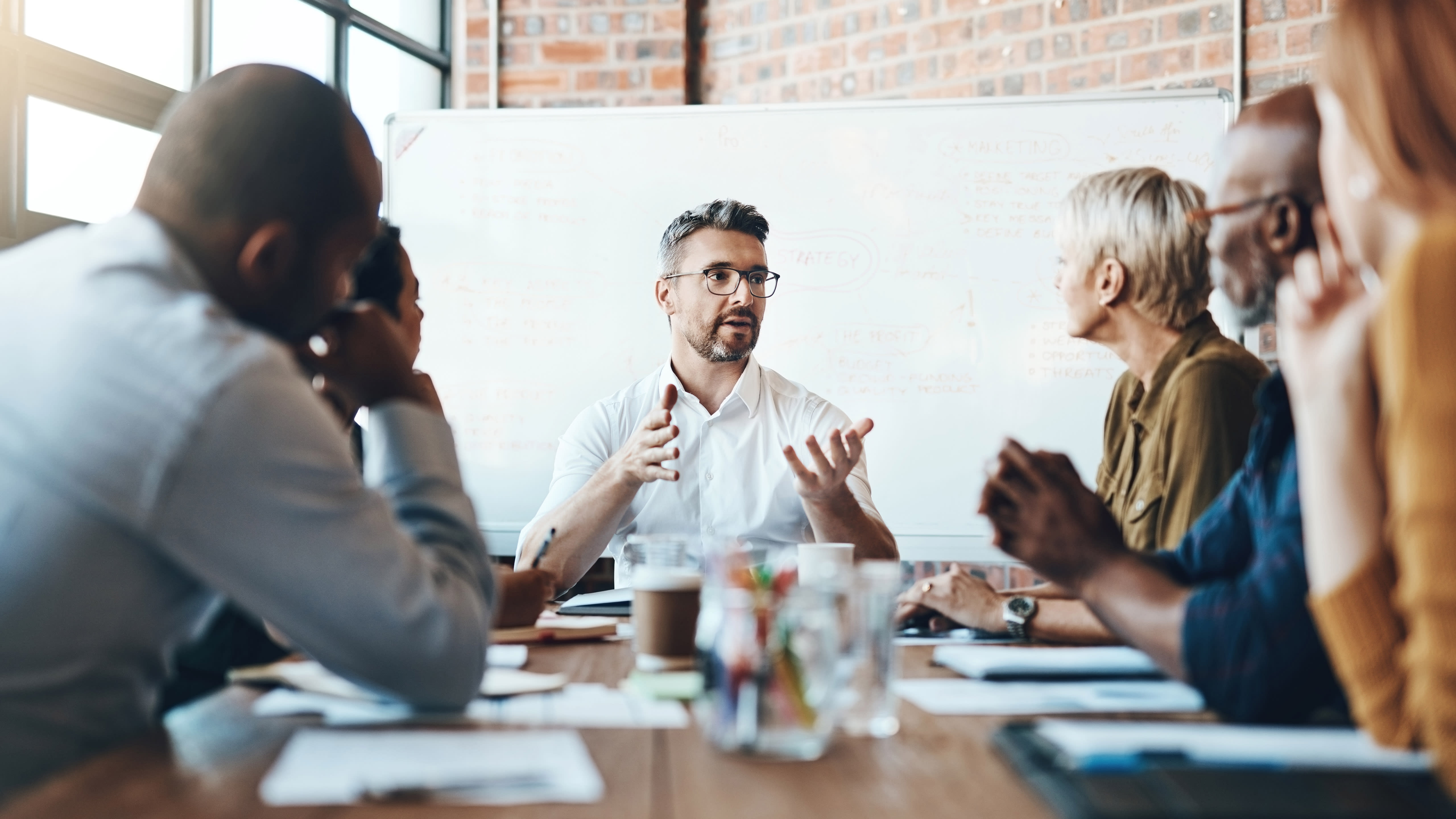 Group of professionals sit at a desk in a brick and glass conference room.