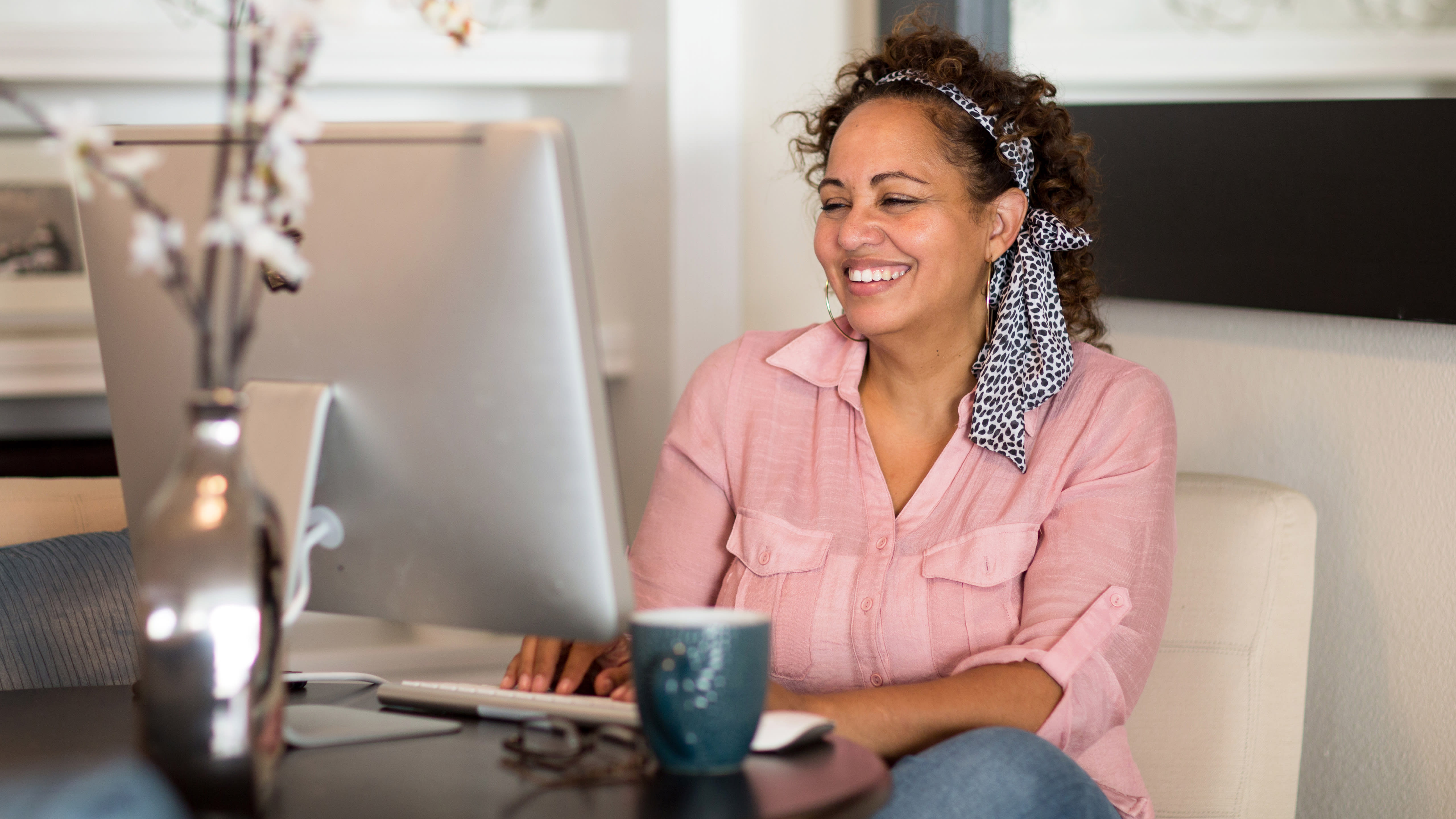 Person sits at desktop computer with coffee mug and decorative flowers in a vase.