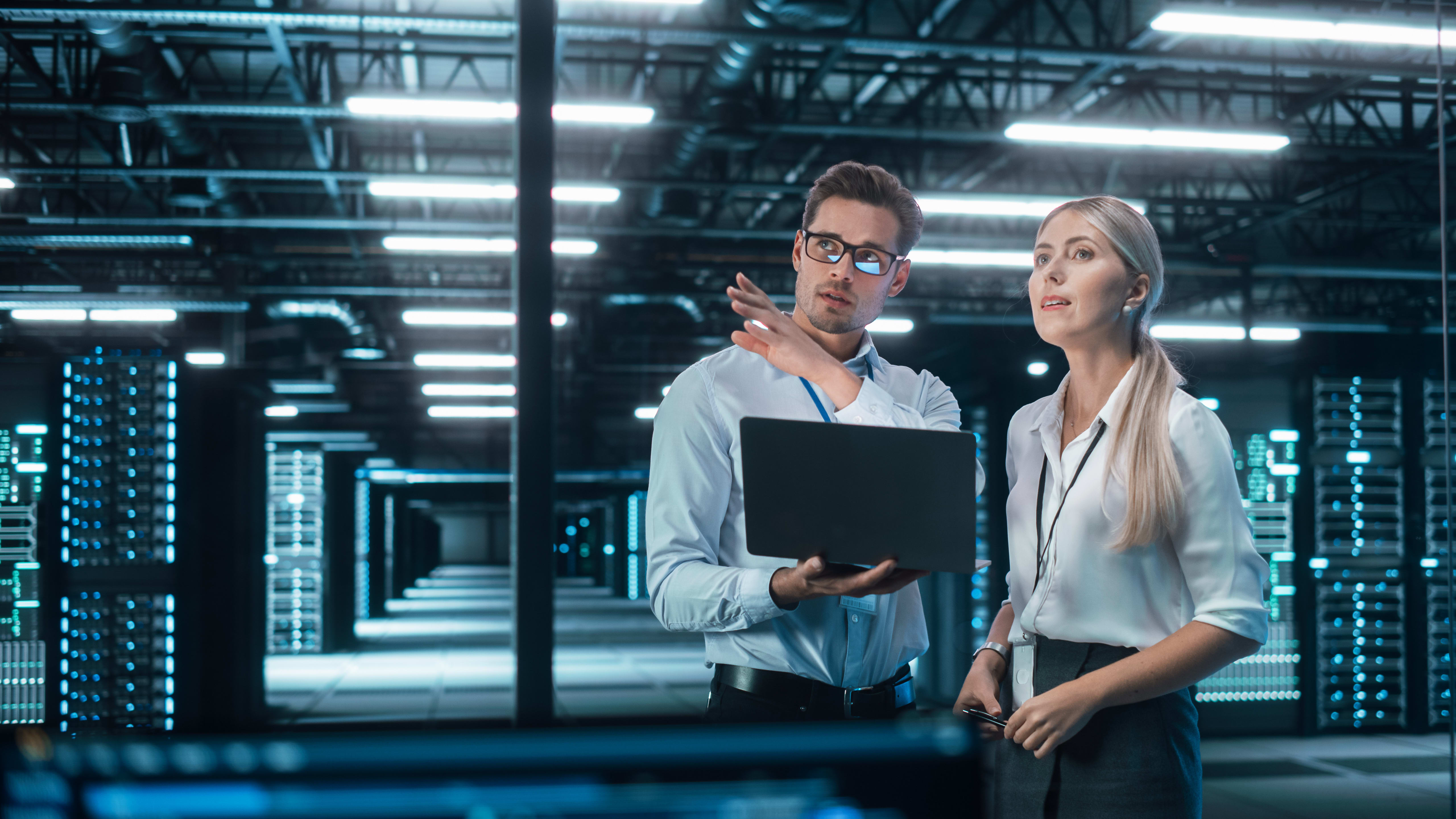 Two colleagues discuss a project while examining a laptop in a high-tech server room filled with illuminated data servers.