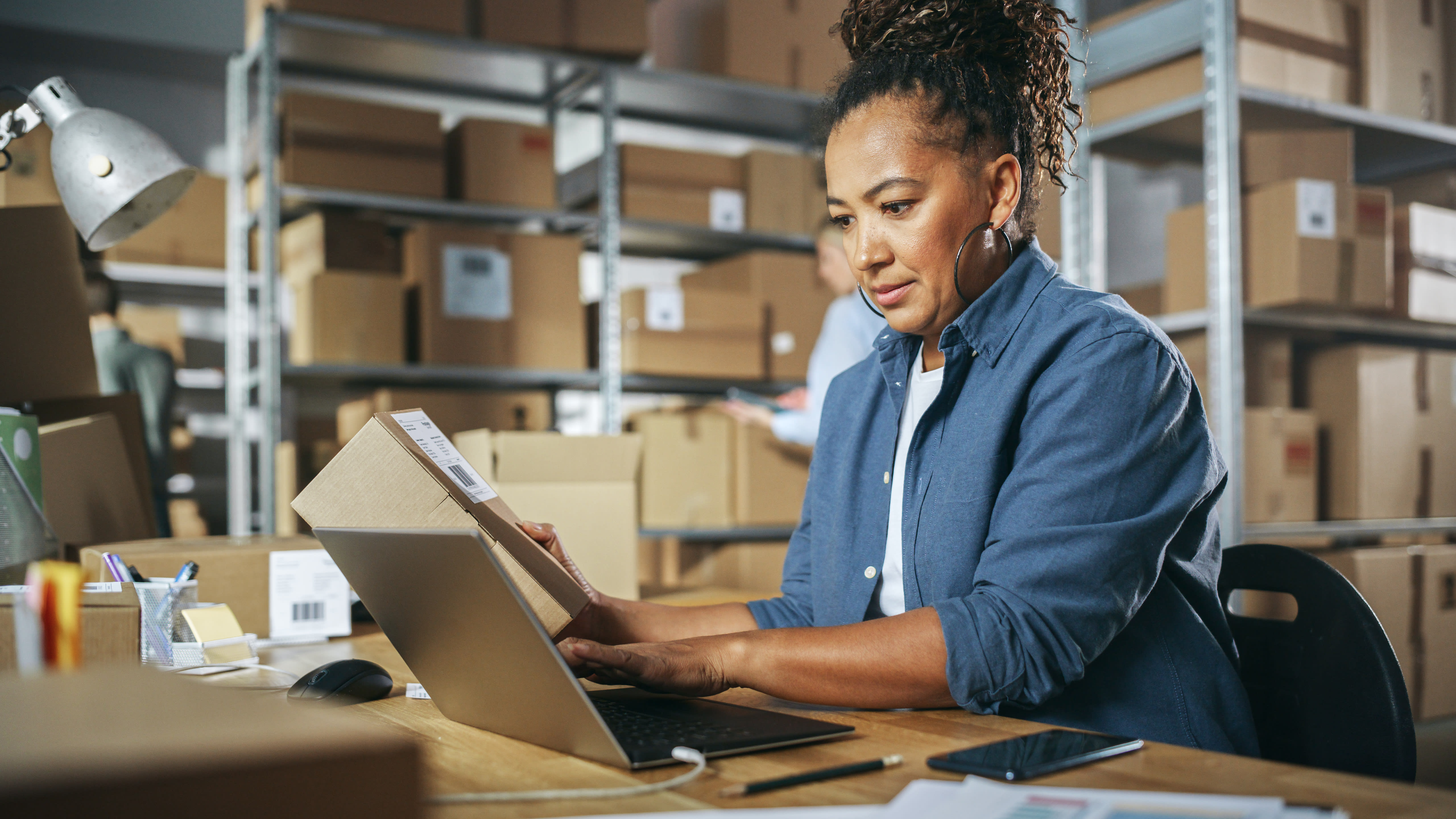 Person types on a laptop while holding paperwork in a warehouse that has stacks of cardboard boxes.