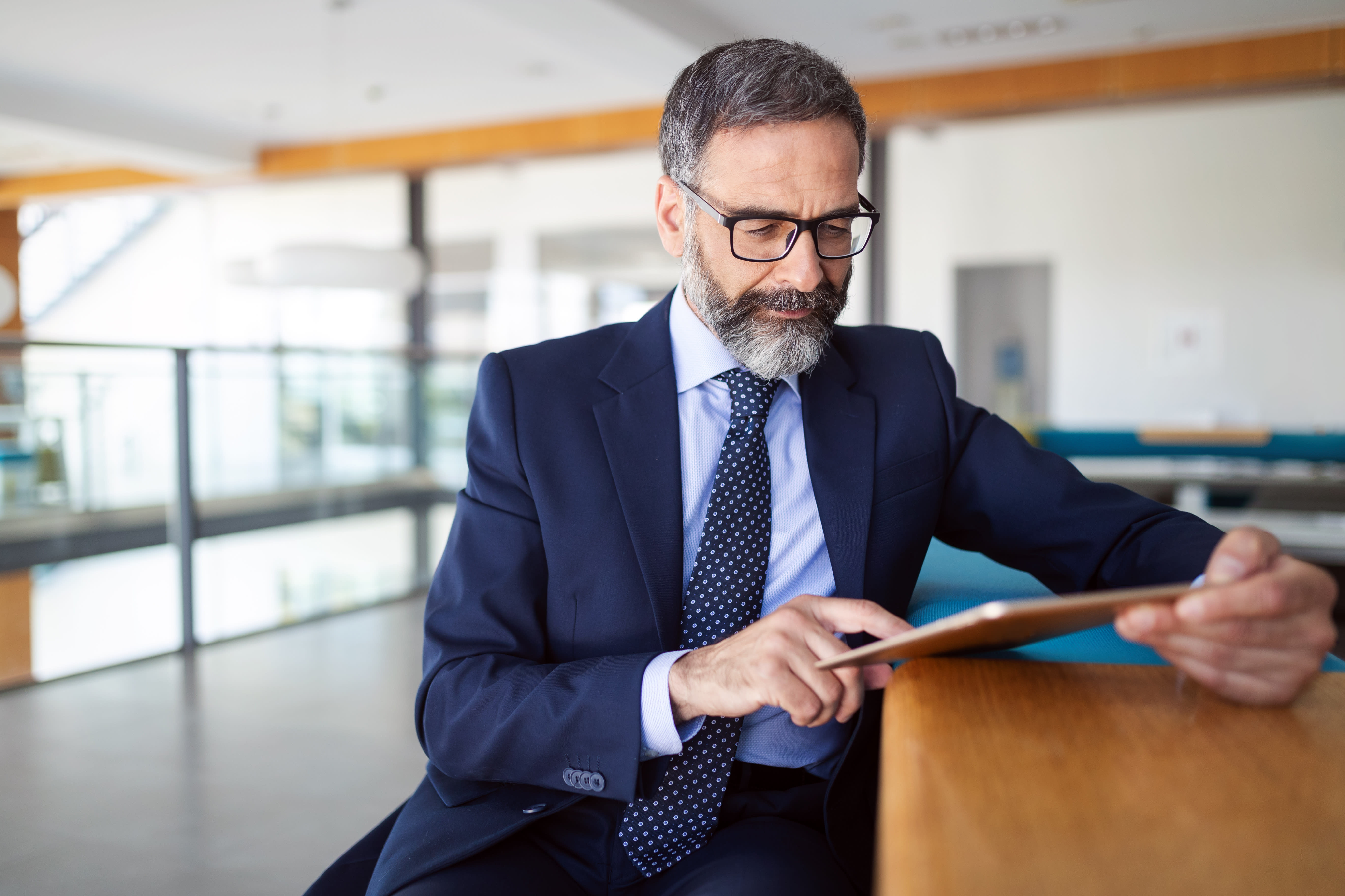 Person wearing a tie and glasses sits at a table and looks at a tablet. 