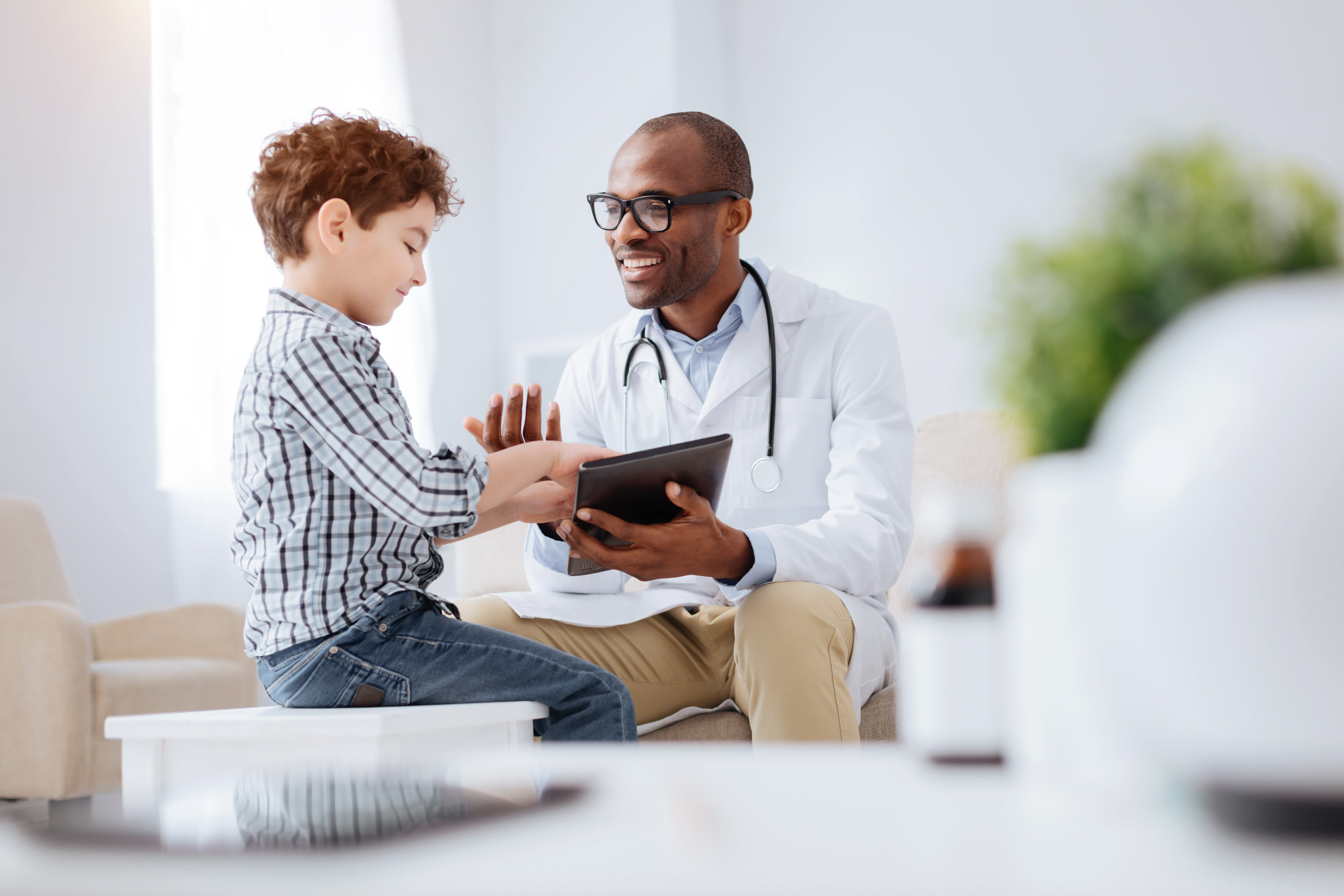 Energetic cheerful male doctor grinning while showing tablet to boy and sitting