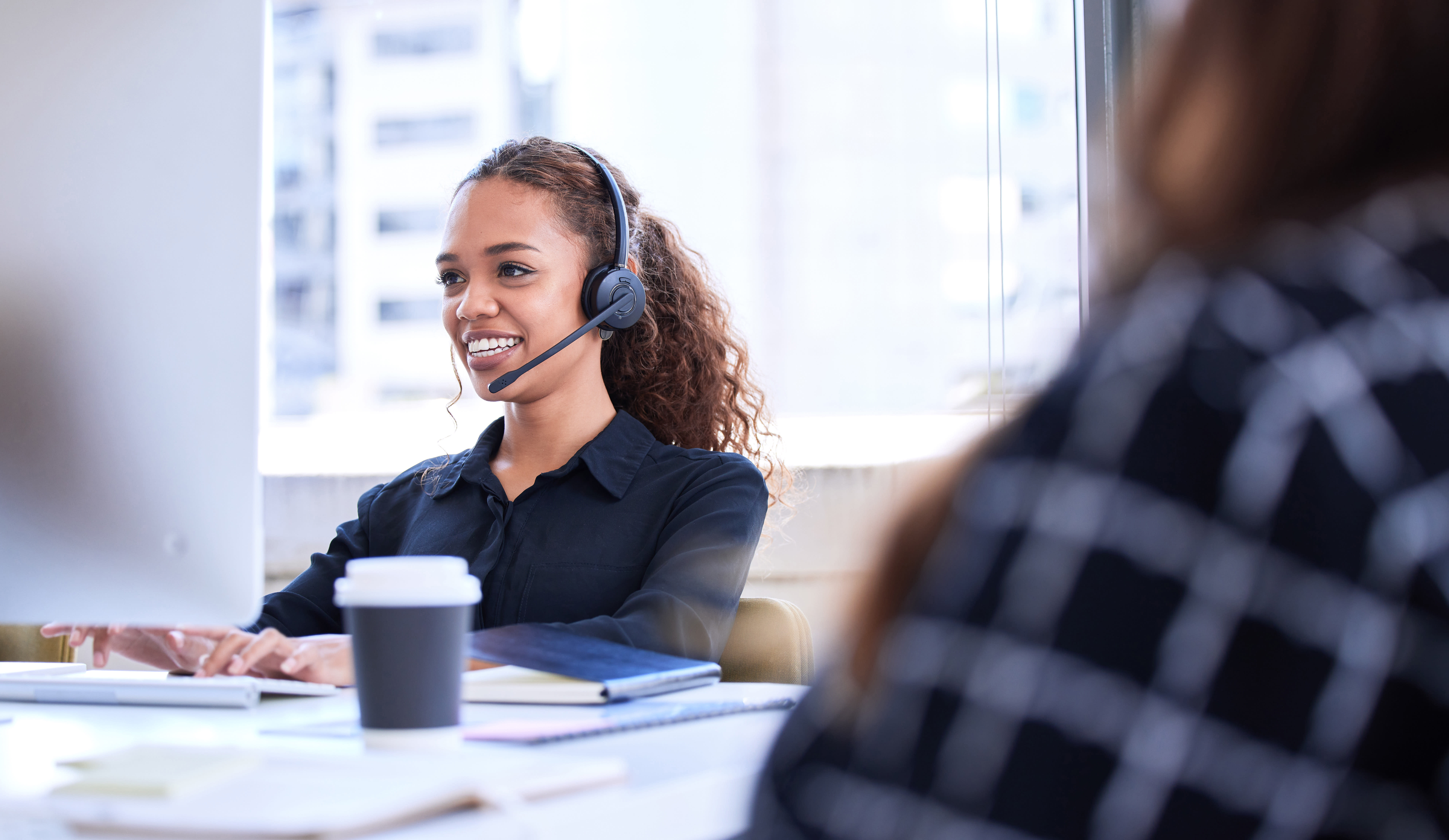 Office employee with a headset works on a desktop with a cup of coffee in the foreground.