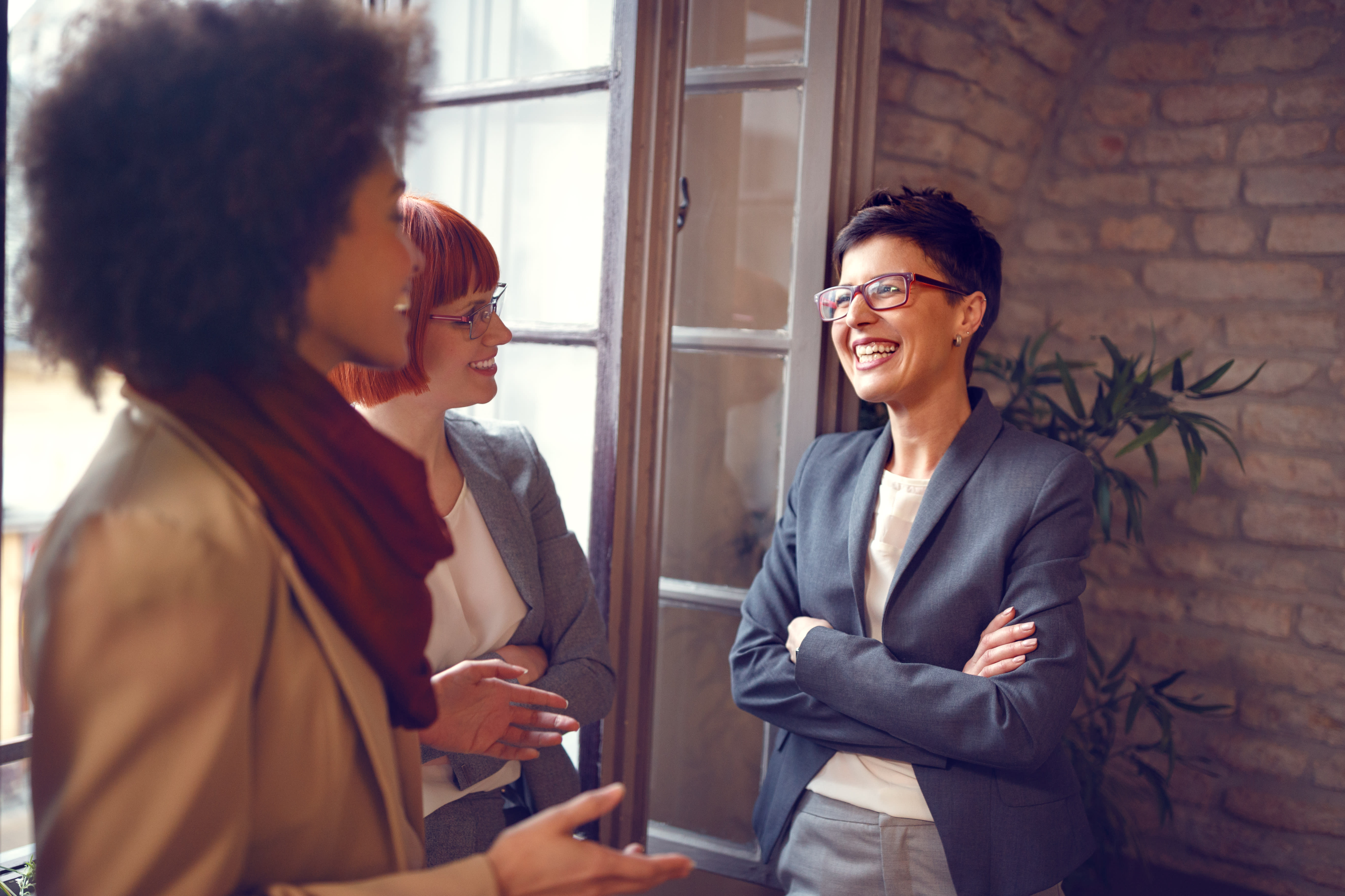 A group of three people in business attire happily chat near a window while working together.