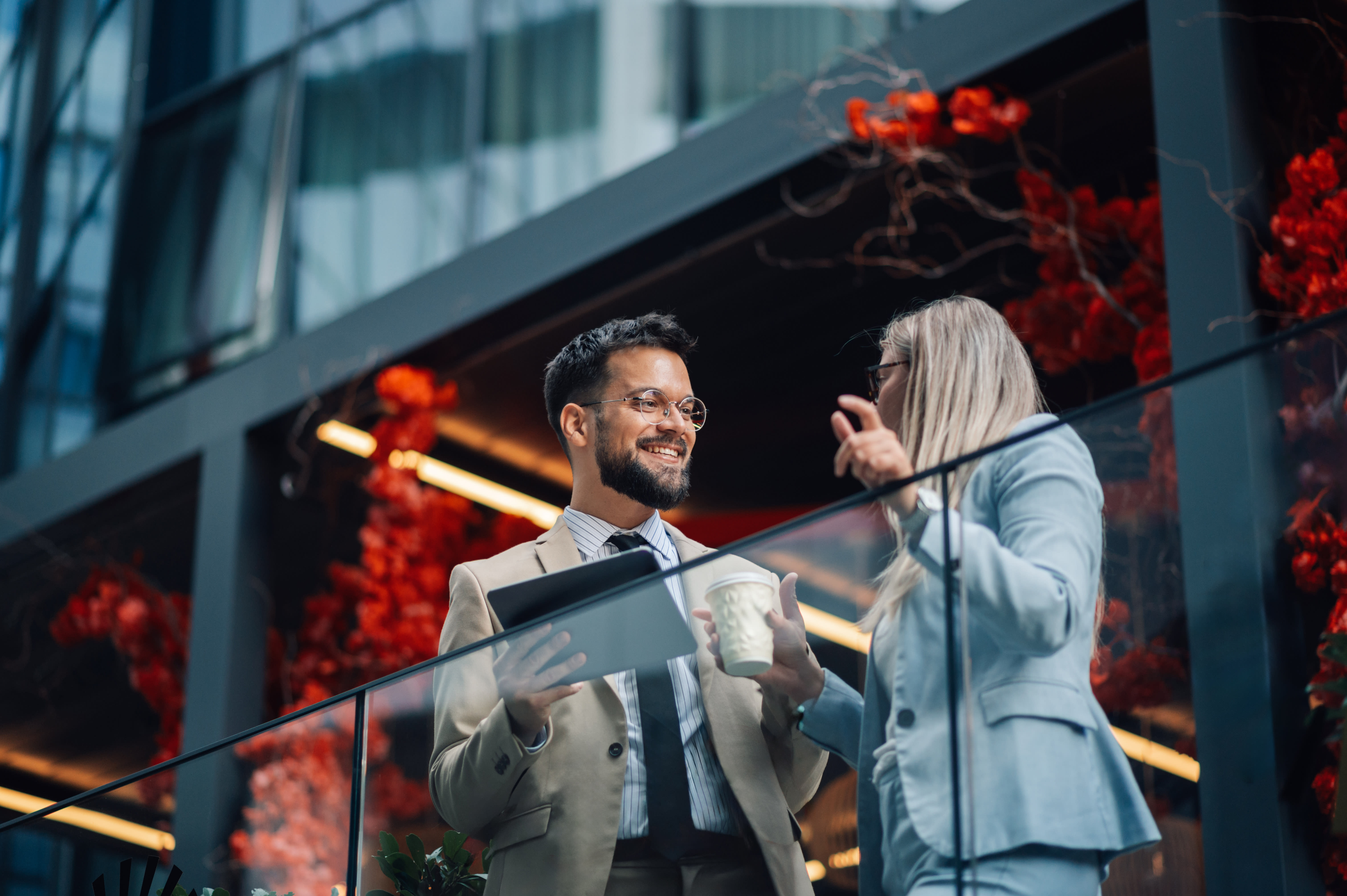 Two young professionals stand and chat outside their office building; a man in a beige suit and glasses holds a tablet while talking to a woman in a gray blazer holding a hot beverage. The leaves on the trees in the background are orange and red, implying that it's fall.