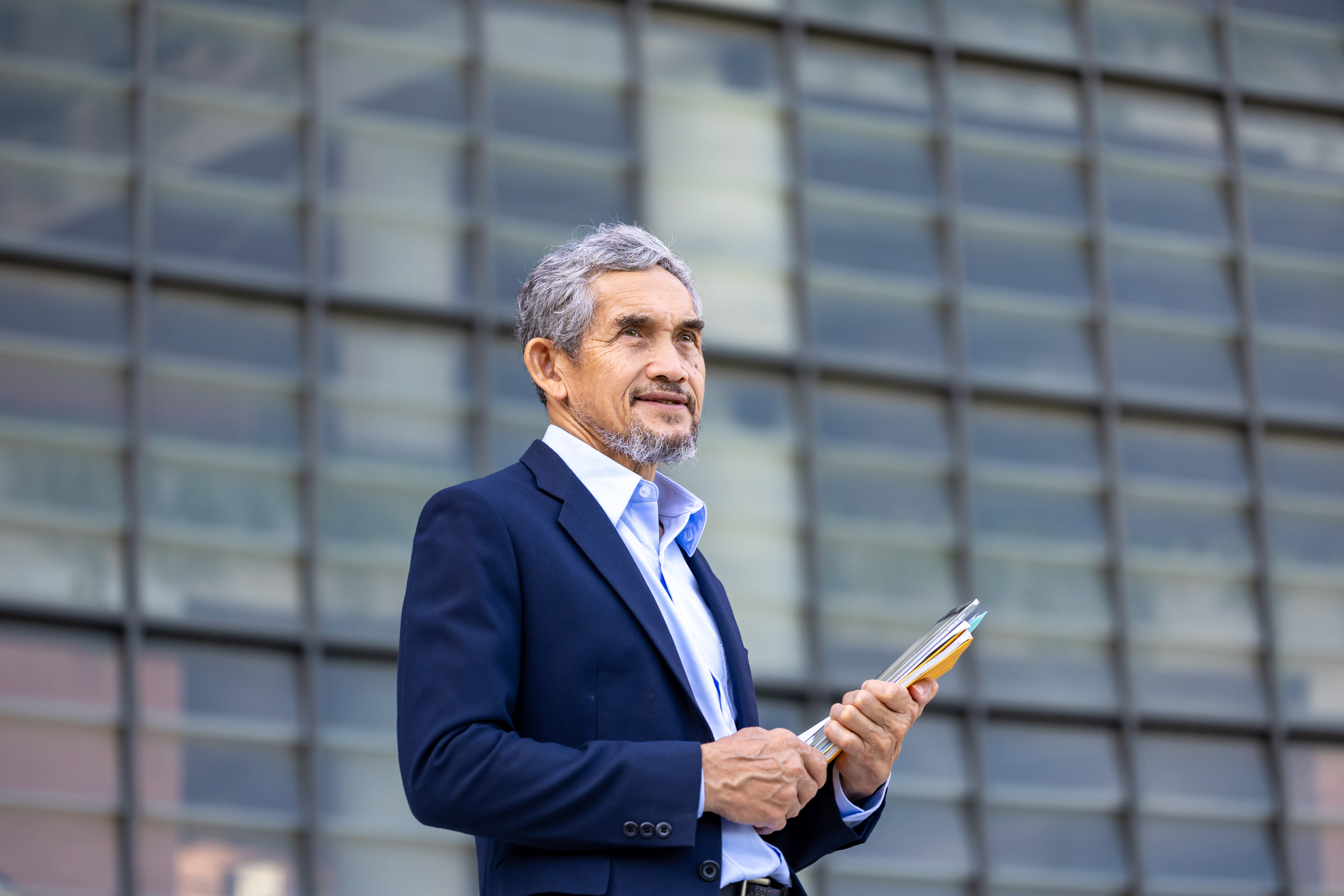 A confident, senior Asian business executive stands outdoors against a cityscape while holding files.