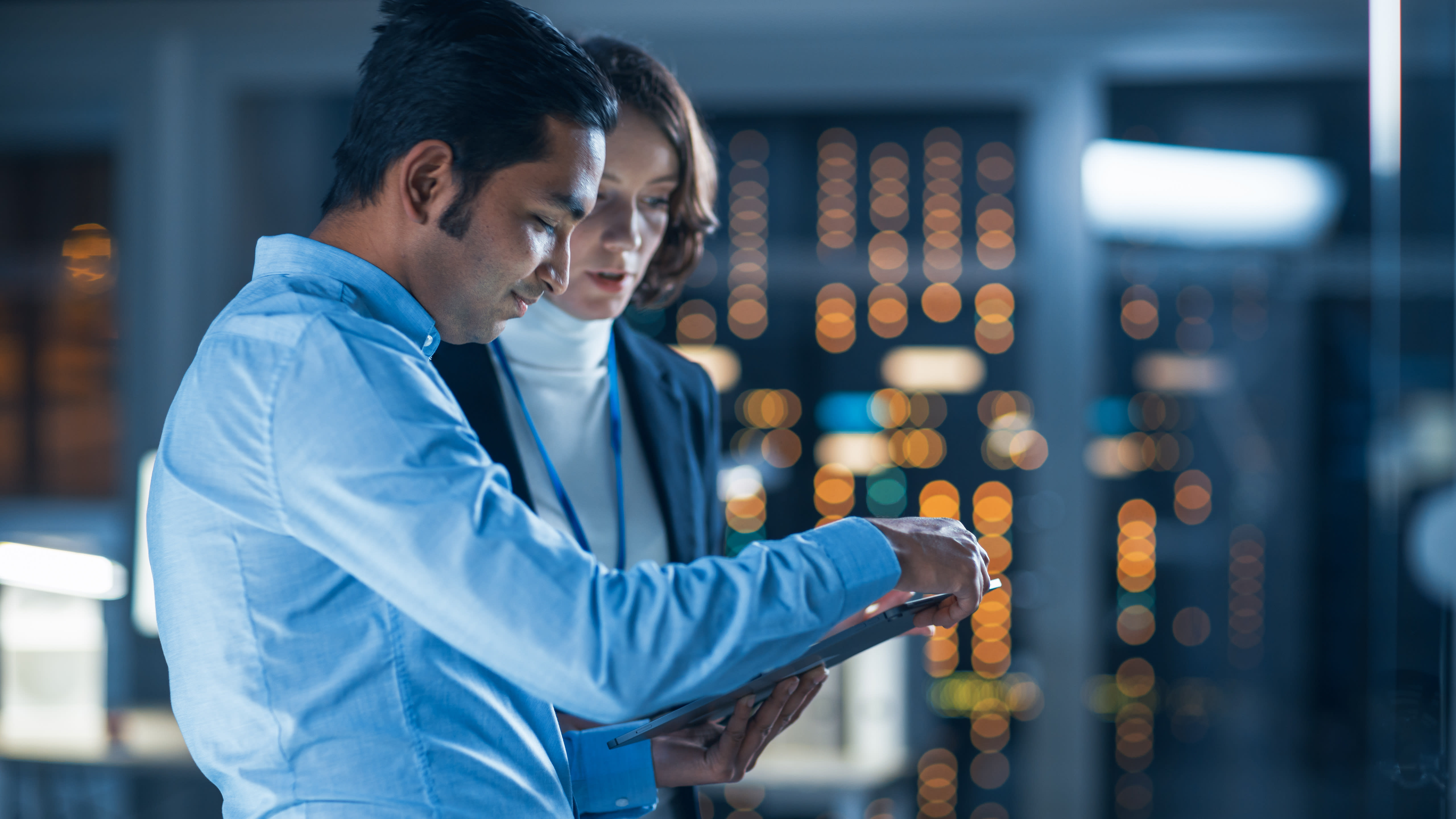 Two professionals, a man and a woman, are collaborating on a tablet in a dimly lit tech server room.