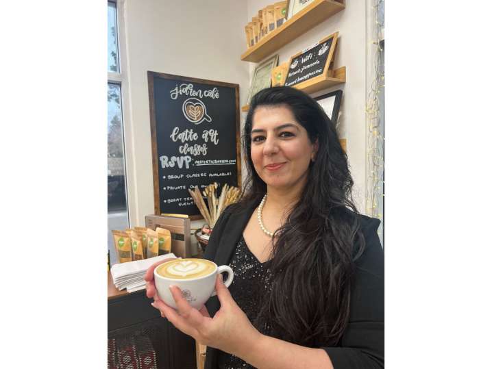 Bay Area - Student holding her latte with a tulip design, smiling in front of a chalkboard that says “coffee with intention.” Shot