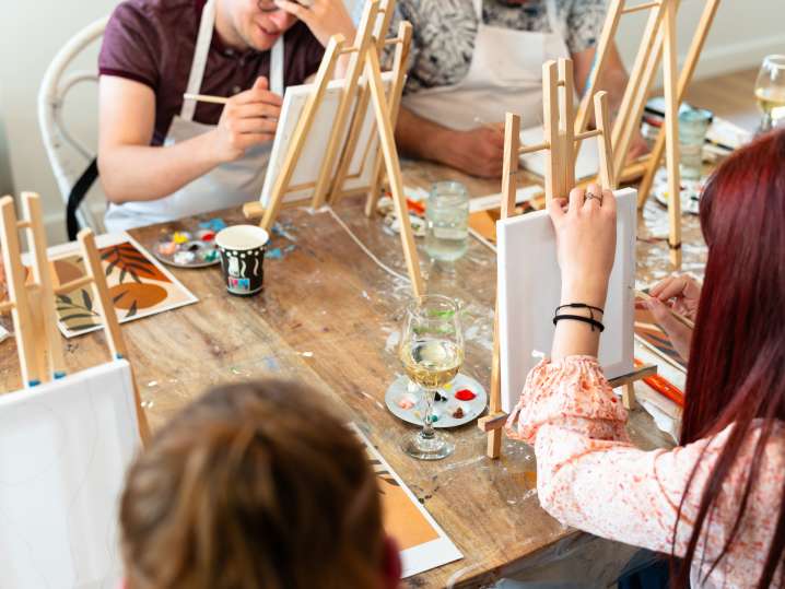 Boston - Overhead view of people painting at a wooden table with drinks and art supplies Shot