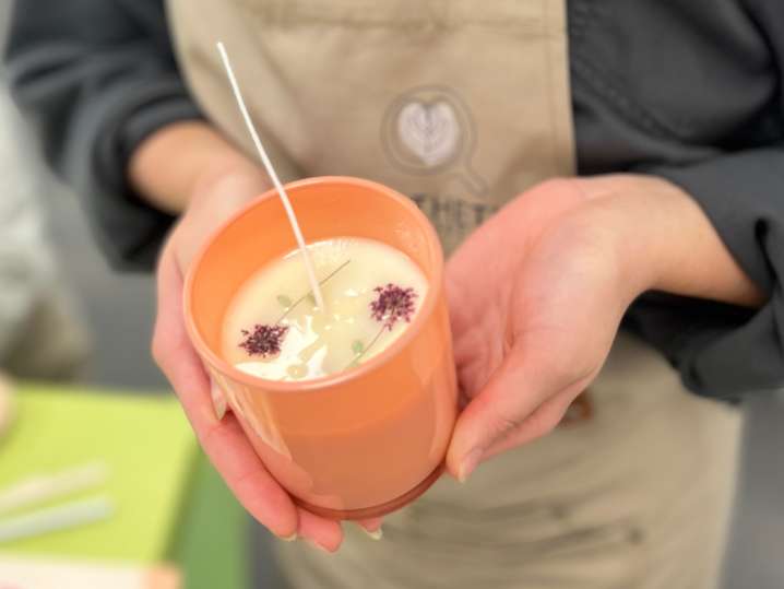 Bay Area - Close-up of a person holding an orange glass filled with creamy wax and dried flower decorations during the candle-making process Shot