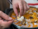 Cooking Class - One Night in Madrid in Downtown Dallas: Close-up of hands placing shrimp and shellfish onto a paella pan filled with golden rice and other seafood.