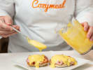 Cooking Class - Eggs Benedict With Finesse in Houston: A chef in a 'Cozymeal' apron pouring hollandaise sauce over Eggs Benedict on a white plate