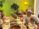 Team Building Cooking Class - Classic Puerto Rican Cooking in Manhattan: A chef in a white coat lifts the lid off a large pot of stew at the center of a long wooden dining table, surrounded by guests in aprons eagerly awaiting the meal