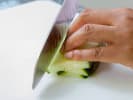 Cooking Class - Vegan Italian Favorites in Boulder: A chef's knife slicing a cucumber into thin planks on a white cutting board, with a hand holding the cucumber steady
