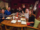 Cooking Class - Cucina Italiana: A Simple Menu: A group of six people sit around a rectangular wooden dining table in a cozy, warmly lit indoor dining area, enjoying a meal together
