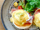Cooking Class - Eggs Benedict With Finesse in Houston: A plated serving of Eggs Benedict with poached eggs, ham, English muffins, and hollandaise sauce, accompanied by a side salad