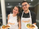 Cooking Class - Simple and Fresh Mediterranean: A smiling man and woman stand side by side in a modern kitchen, each wearing a white apron with the orange Cozymeal logo