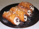 Cooking Class - Authentic Pizza and Italian Cuisine: Four golden brown cannoli filled with creamy ricotta and topped with mini chocolate chips, neatly arranged on a black plate and lightly dusted with powdered sugar, placed on a white marble counter