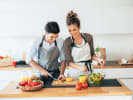 Cooking Class - Cook a Delicious Mediterranean Feast - Date Night in Downtown Dallas: a couple cooking Mediterranean dishes together in the kitchen while smiling and wearing aprons