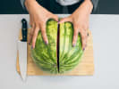 Cooking Class - Garden Gourmet in Manhattan: Cutting a whole watermelon in half â€“ Overhead view of hands slicing a whole watermelon in half on a wooden cutting board with a chefâ€™s knife beside it.