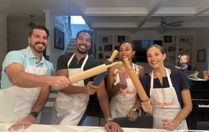 Four participants wearing Cozymeal aprons hold rolling pins behind a kitchen counter during a pasta-making class in Houston.