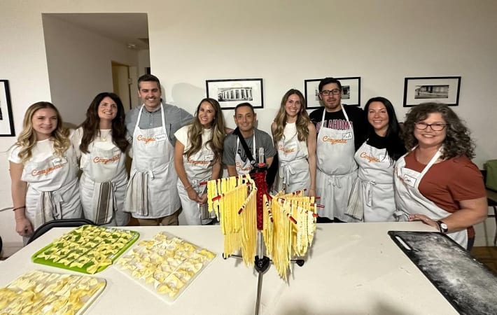 Nine participants wearing Cozymeal aprons stand behind trays of handmade pasta and pasta drying racks in a pasta-making class in NYC.
