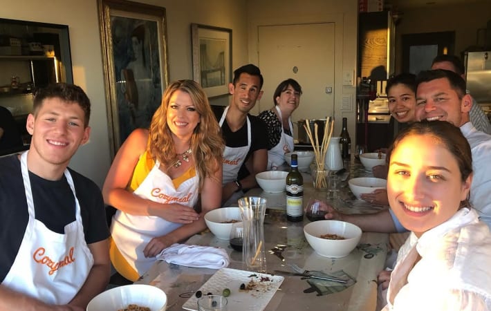 Participants wearing Cozymeal aprons sit around a table with bowls of prepared food during a team building activity in Houston.