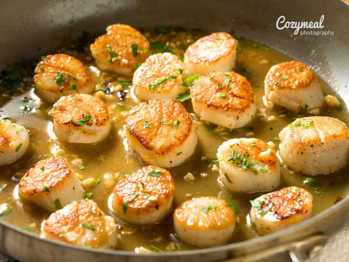 Seared scallops â€“ Golden-brown seared scallops garnished with herbs, sizzling in a pan with melted butter.