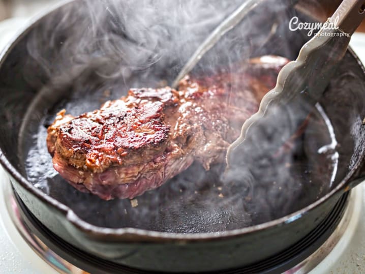 Seared steak cooking in a cast iron skillet with smoke rising and tongs holding the meat.