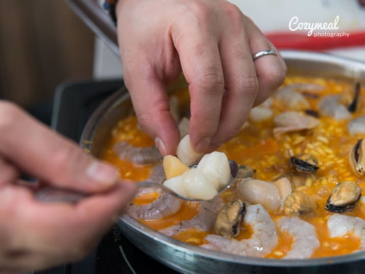 Close-up of hands placing shrimp and shellfish onto a paella pan filled with golden rice and other seafood.