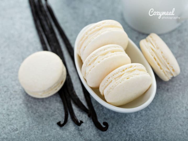 White bowl of vanilla macarons with whole vanilla beans on a gray background.