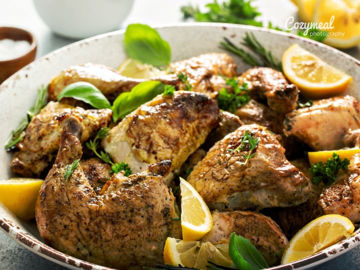 Golden roasted chicken pieces served in a white dish, garnished with fresh herbs and lemon wedges.