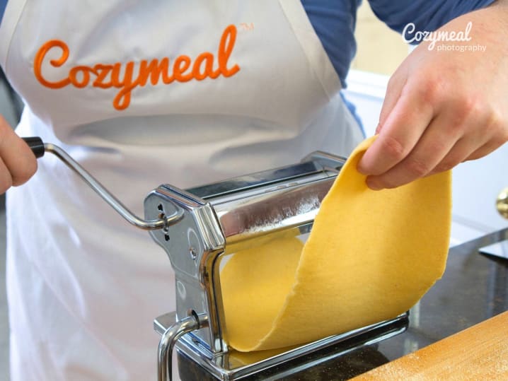 Rolling pasta dough through a machine with a Cozymeal apron visible.