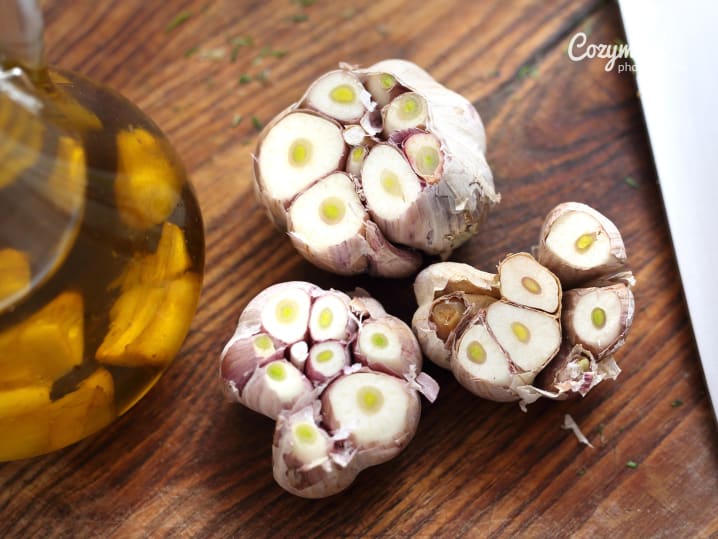 Close-up of halved garlic bulbs on a rustic wooden board beside a glass bottle of olive oil.