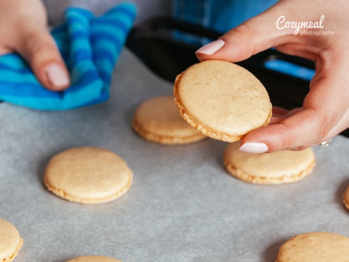 Hands sandwiching vanilla macaron shells together with buttercream filling.