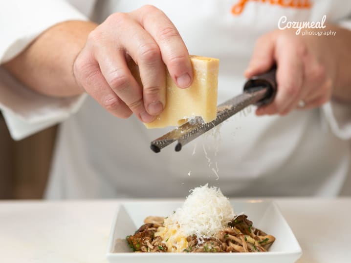 Hand grating cheese over risotto on a white plate, garnished with herbs.