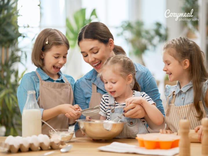 A family of kids and an adult gathered around a bowl, baking together at a table.