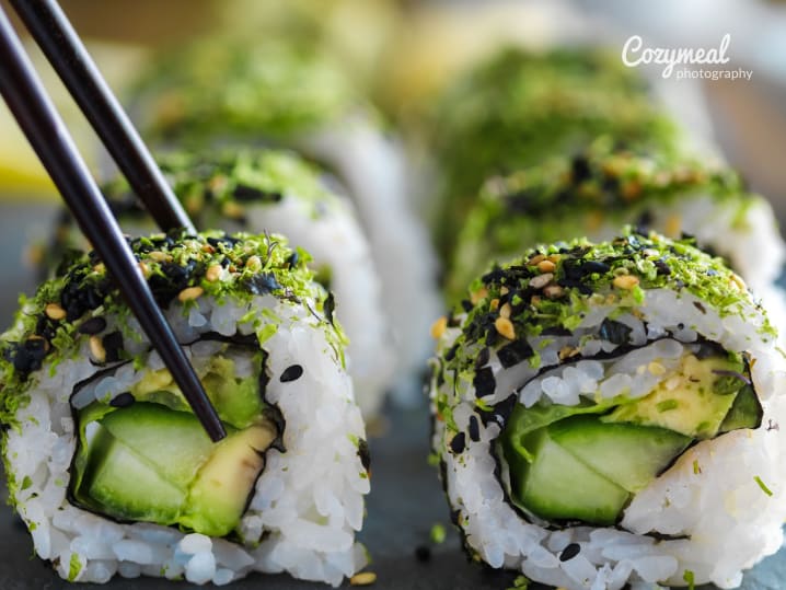 Close-up of avocado and vegetable sushi rolls topped with sesame seeds, with chopsticks.